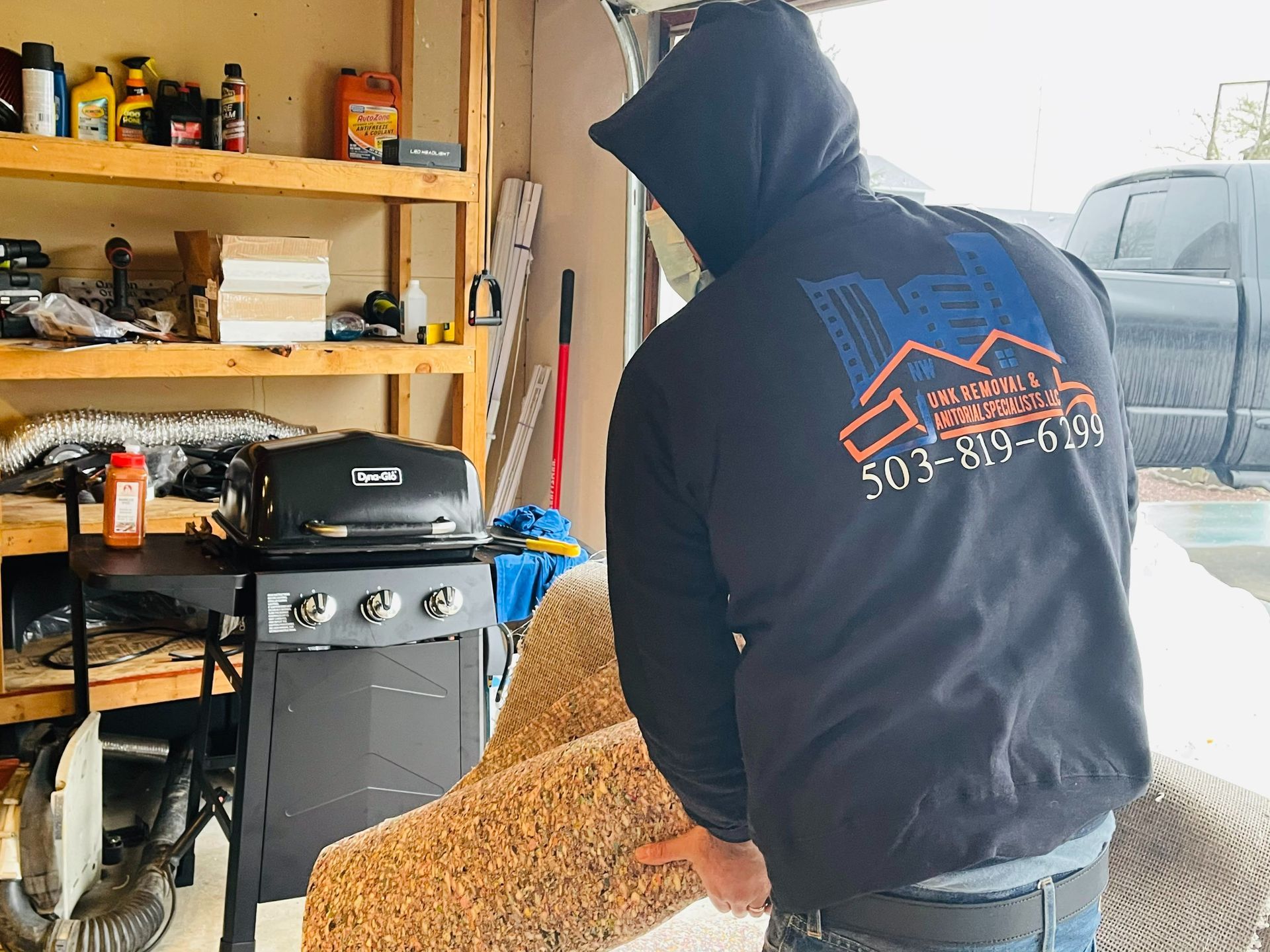 Person in black hoodie carrying a tan-colored rolled rug in a garage, next to a grill and shelving.