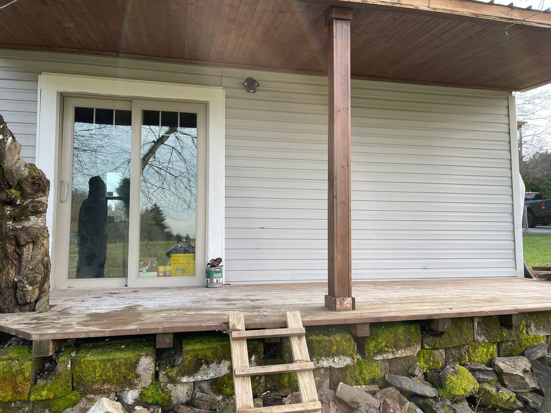 A building with a sliding glass door and brown post on a wooden deck over a mossy stone wall.
