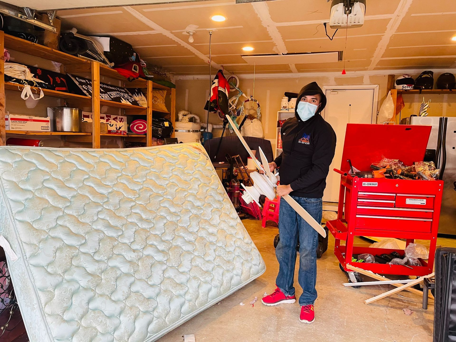 Man in mask holding wooden stick in a messy garage next to a mattress, possibly breaking it.
