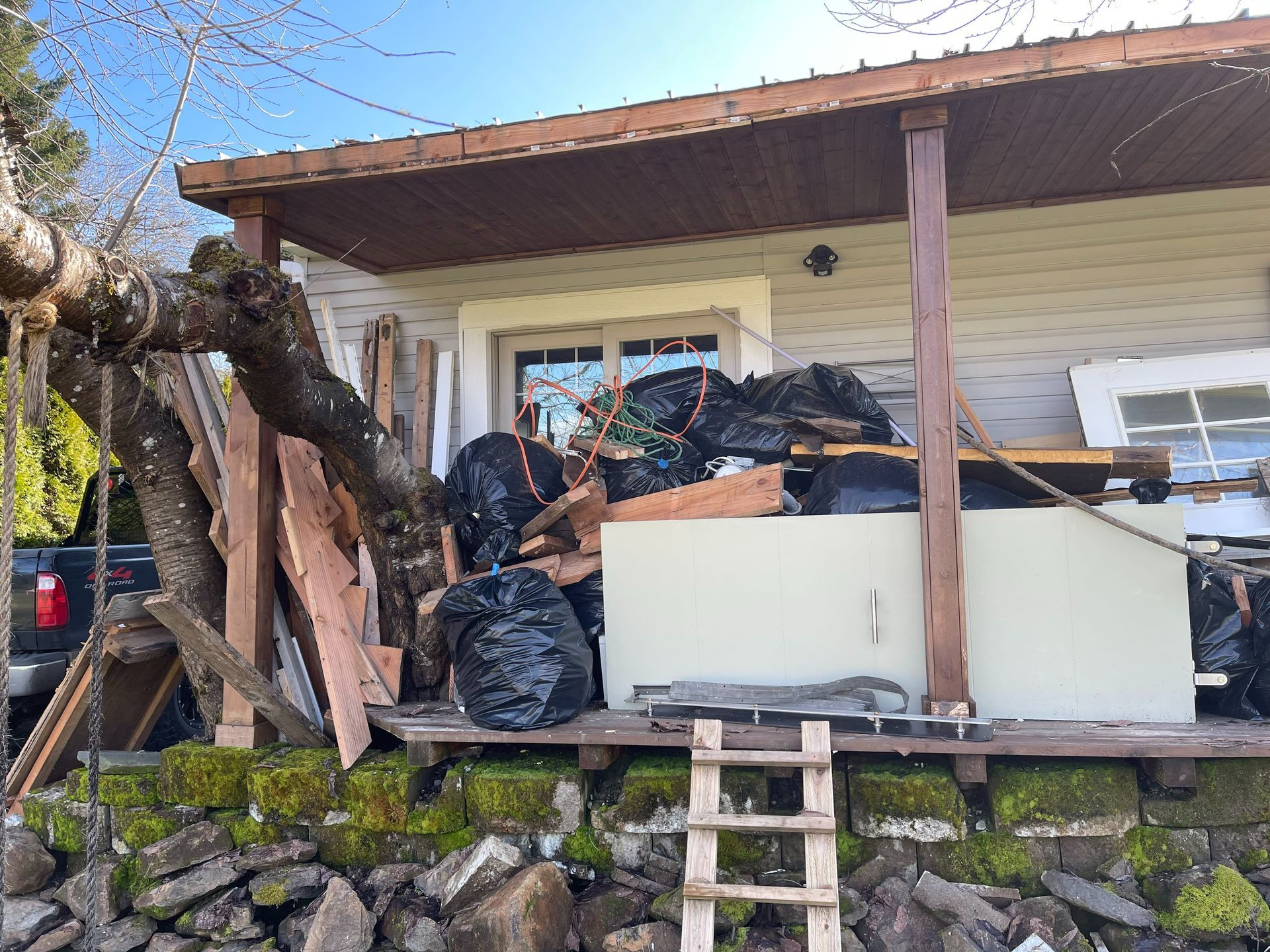 A cluttered porch with black trash bags, wood, and a small refrigerator. A ladder leads down to a stone wall.