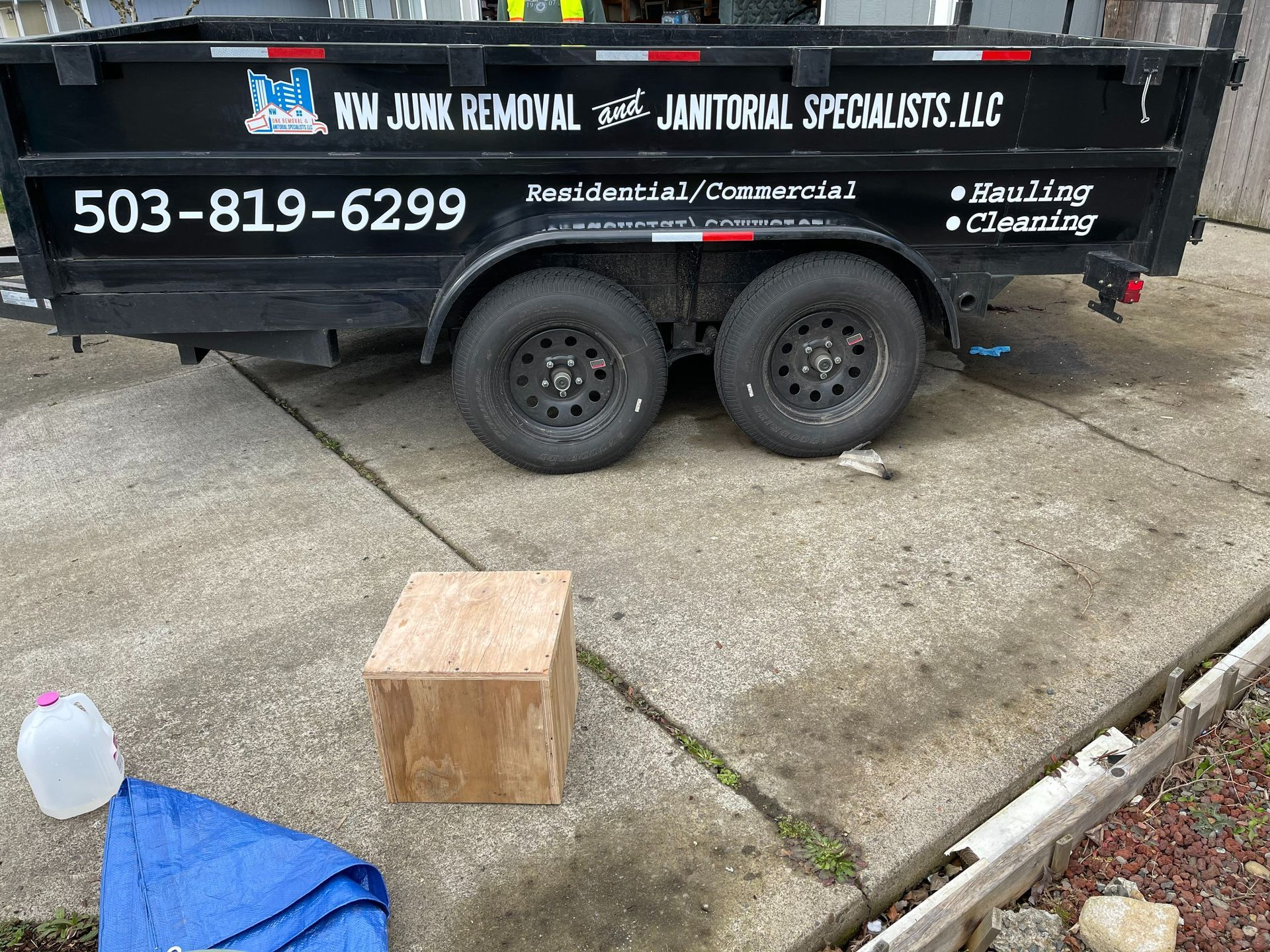 Black trailer for junk removal, wooden box, water jug, and blue tarp on concrete.