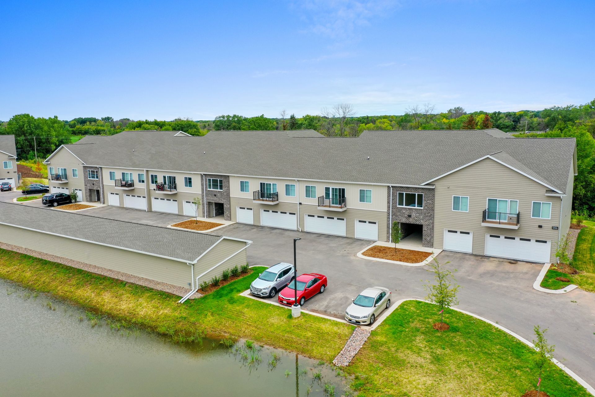 Aerial view of a tan apartment building beside a pond, with parked cars and green lawn.