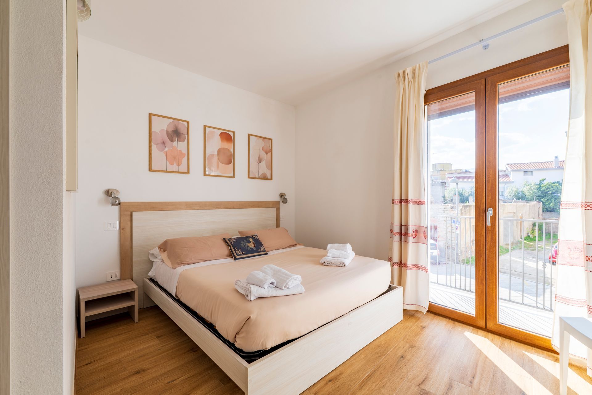 Una camera da letto con letto matrimoniale queen-size, pavimento in legno chiaro, comodino, tre stampe alle pareti e porta a vetri con tende.