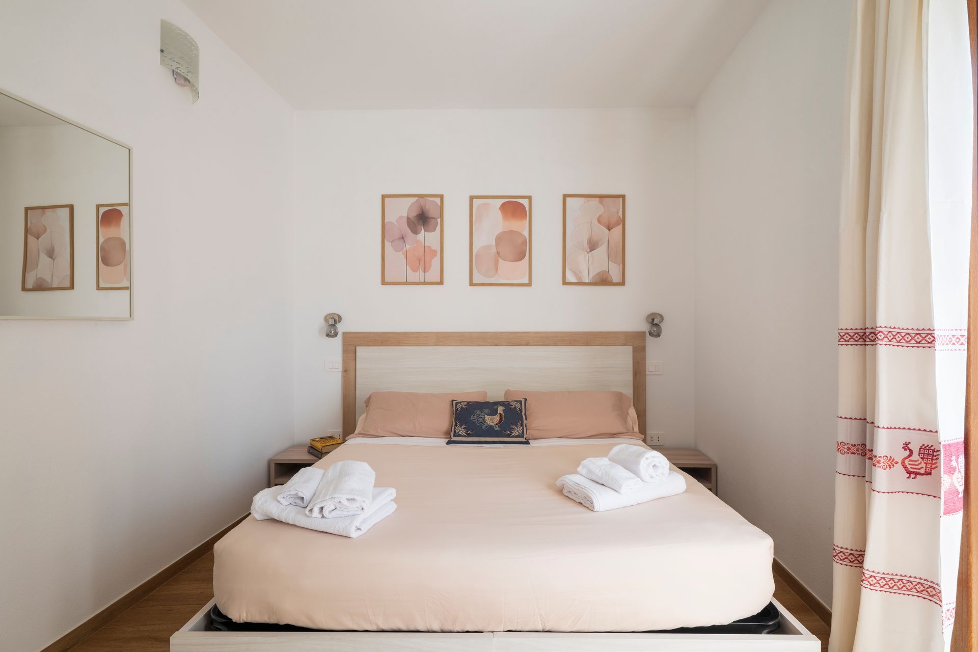 Una camera da letto luminosa con un letto matrimoniale queen size, lenzuola color pesca, asciugamani piegati, tre stampe astratte e tende a fiori.