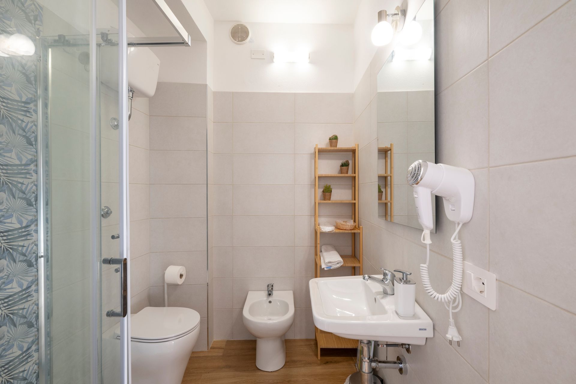 Un bagno luminoso e moderno con doccia in vetro, WC, bidet, lavabo con mobiletto e asciugacapelli a parete.