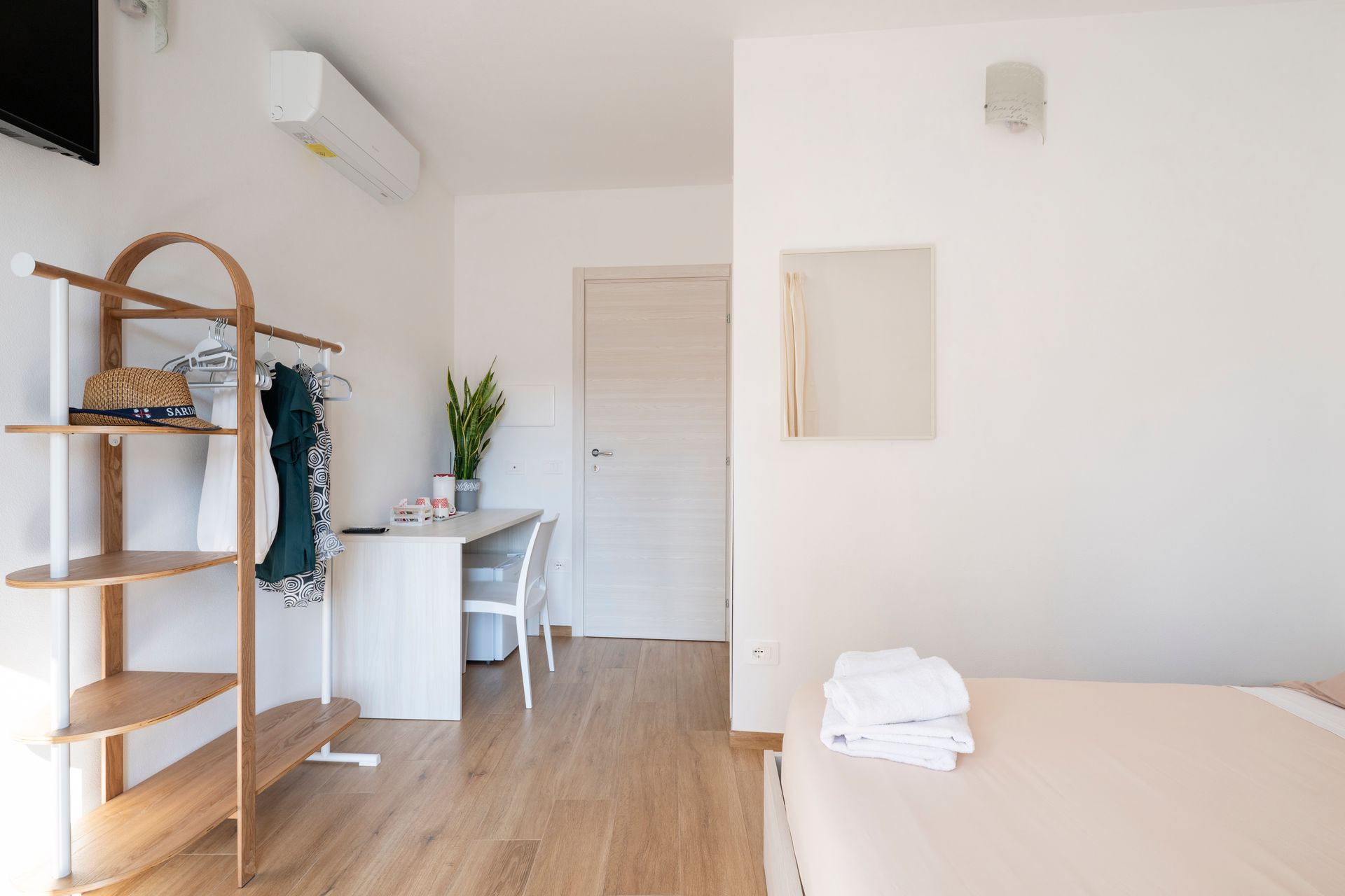 Una camera d'albergo luminosa e minimalista, con un appendiabiti in legno, una scrivania con una pianta e un letto con asciugamani bianchi piegati.