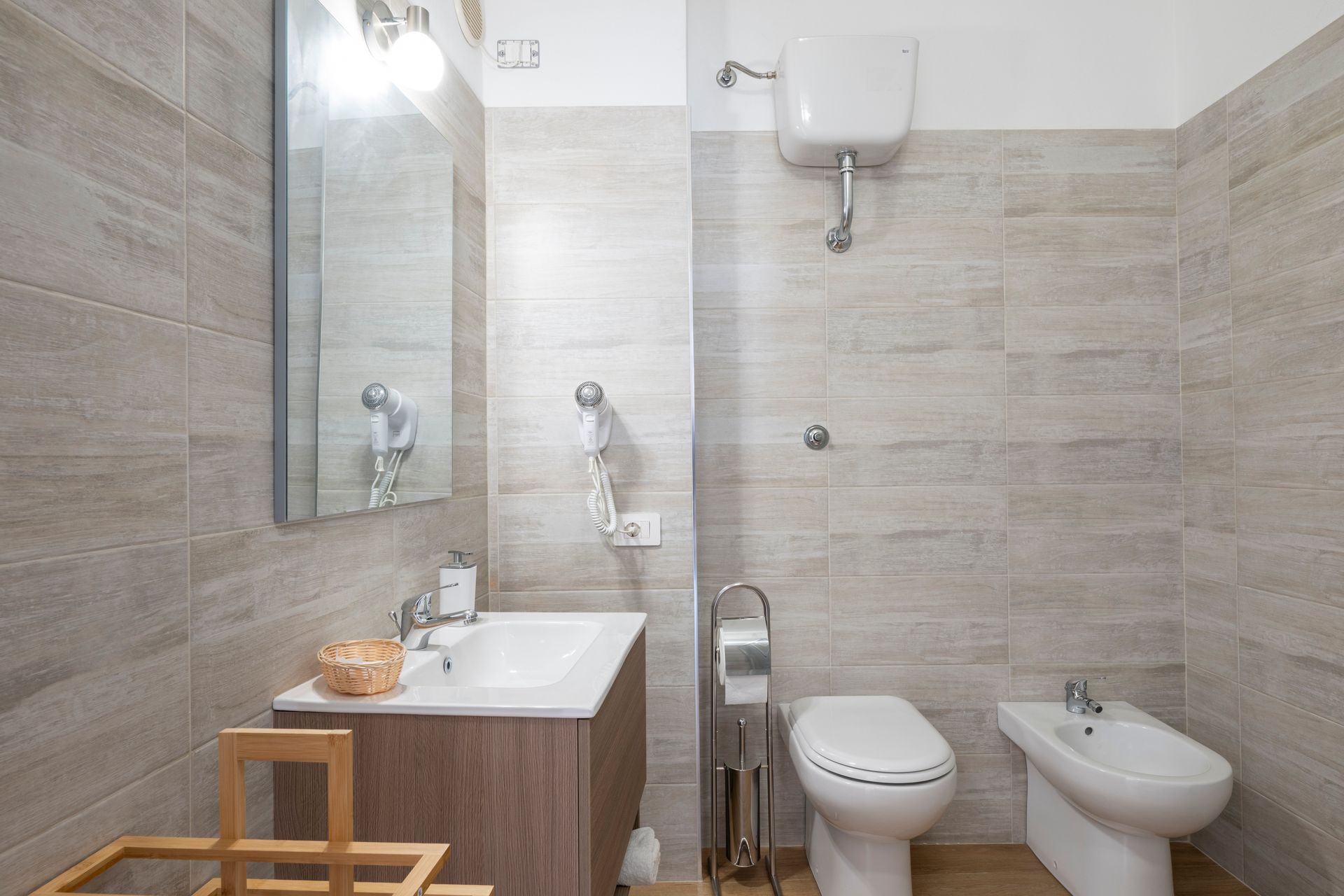 Un bagno con mobiletto in legno dotato di lavabo, specchio, WC sospeso e bidet, il tutto su pareti rivestite con piastrelle beige.