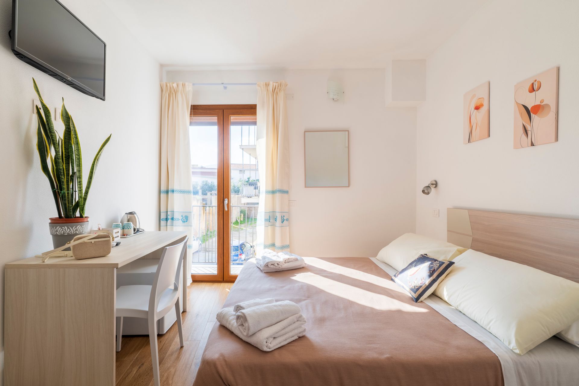 Una camera d'albergo luminosa con un letto con lenzuola bianche, una scrivania con una pianta e una porta a vetri che conduce a un balcone.