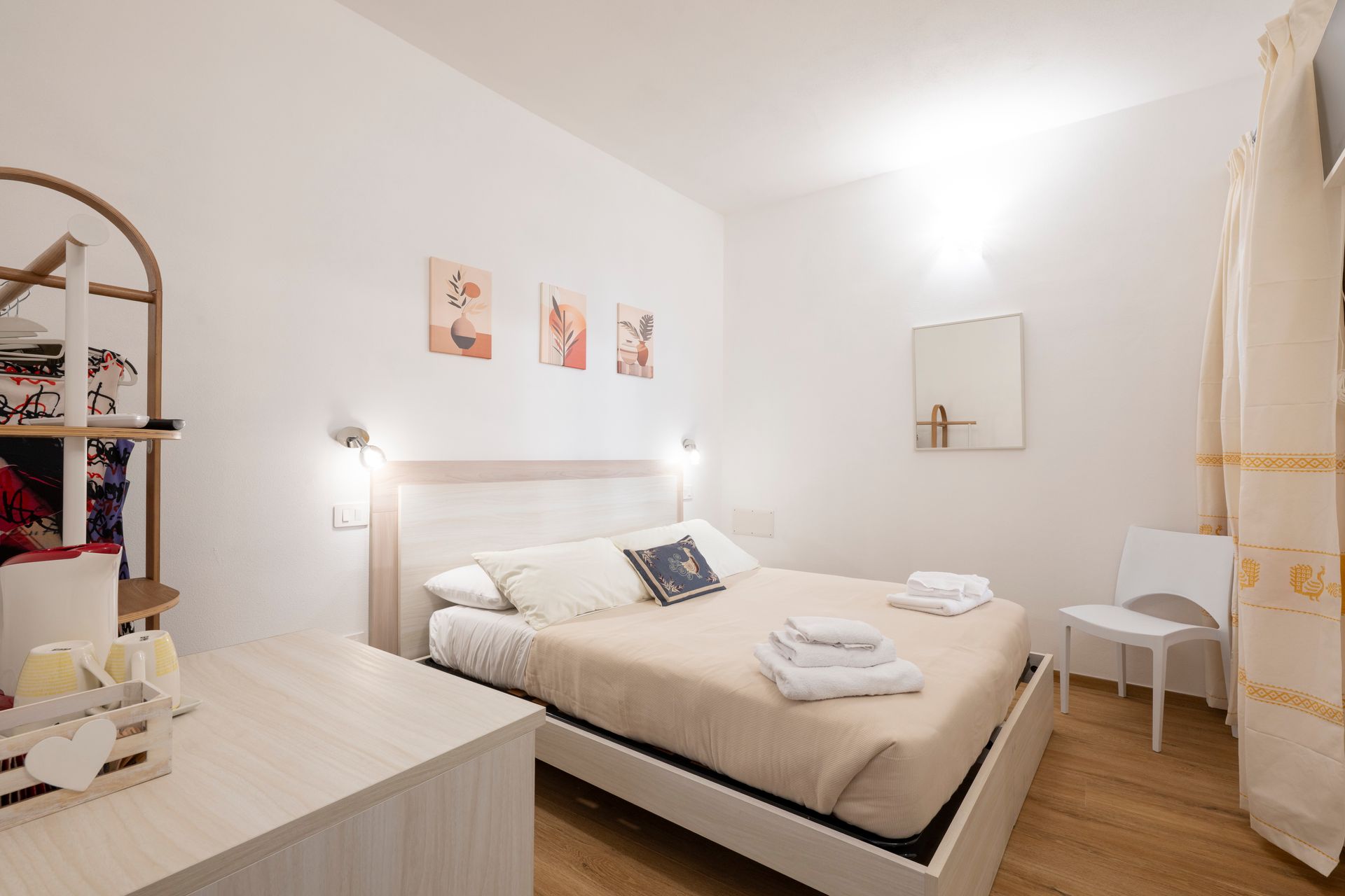 Una camera da letto con un letto matrimoniale con lenzuola beige, tre piccoli quadri alle pareti, una sedia bianca e uno scaffale in legno.