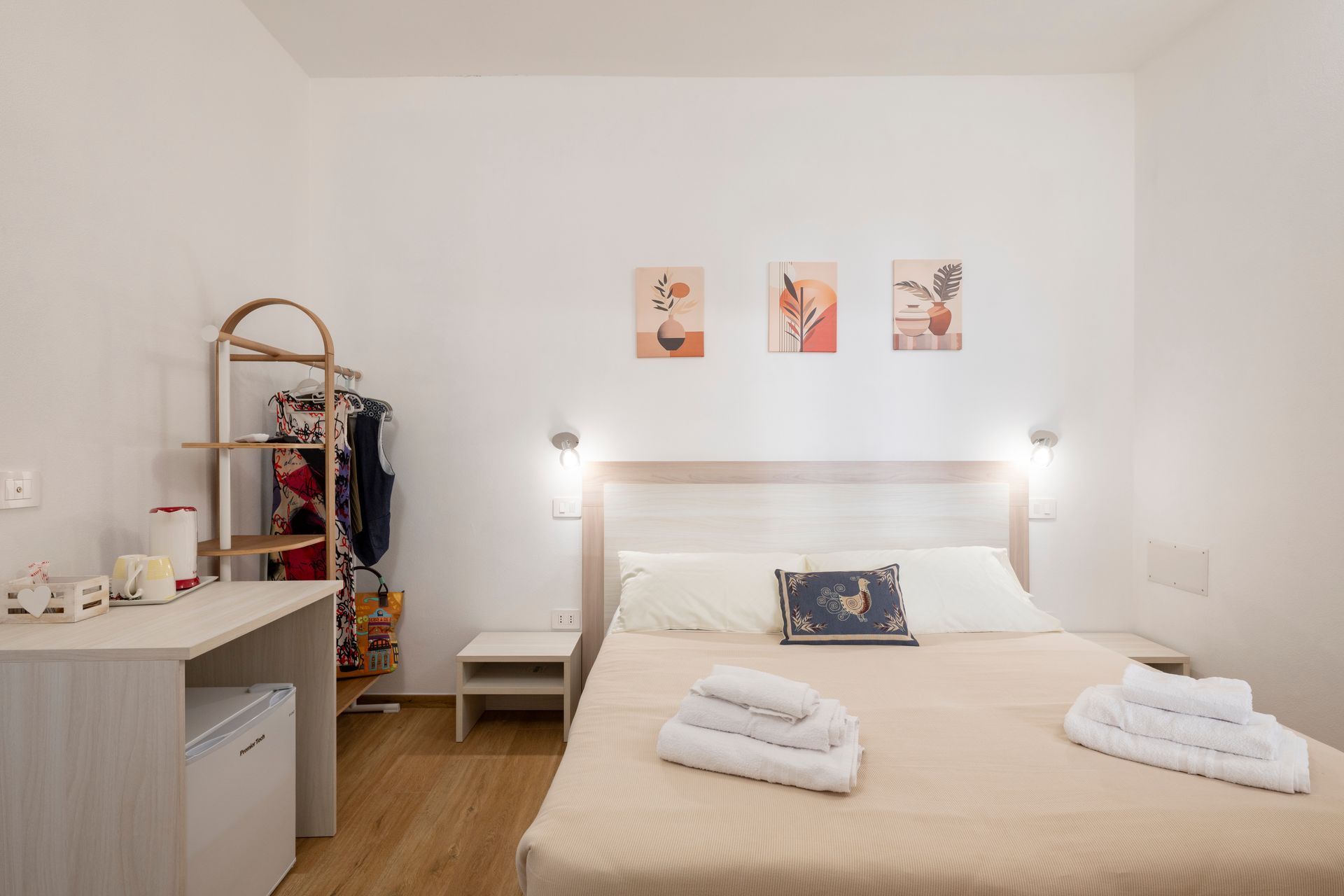 Una camera da letto moderna e minimalista con letto matrimoniale, mobili in legno chiaro, un appendiabiti e tre stampe alle pareti.