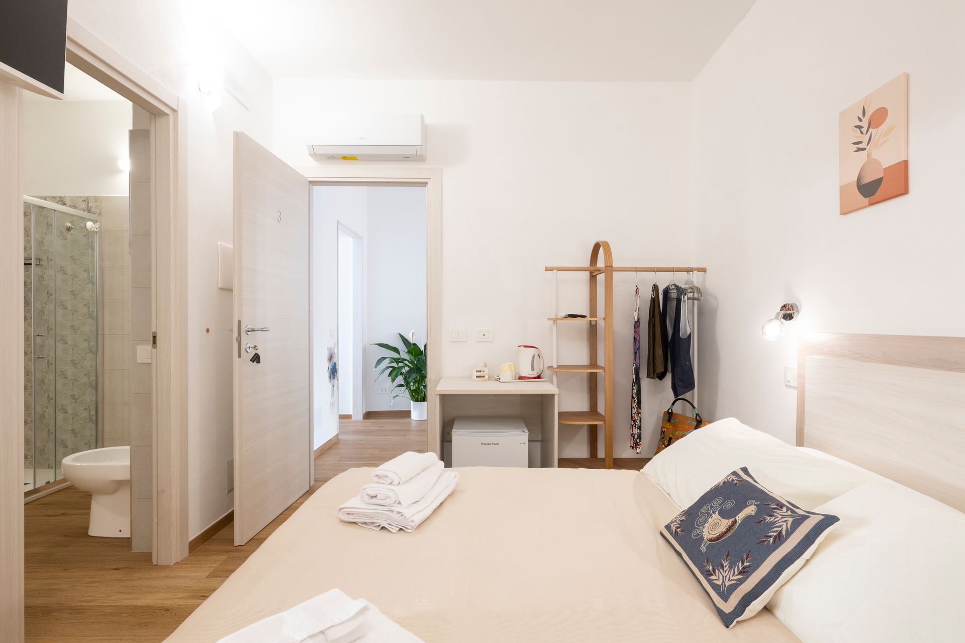 Una moderna camera d'albergo con un letto ben rifatto, un piccolo armadio a giorno, un bagno privato e arredi dai colori chiari.