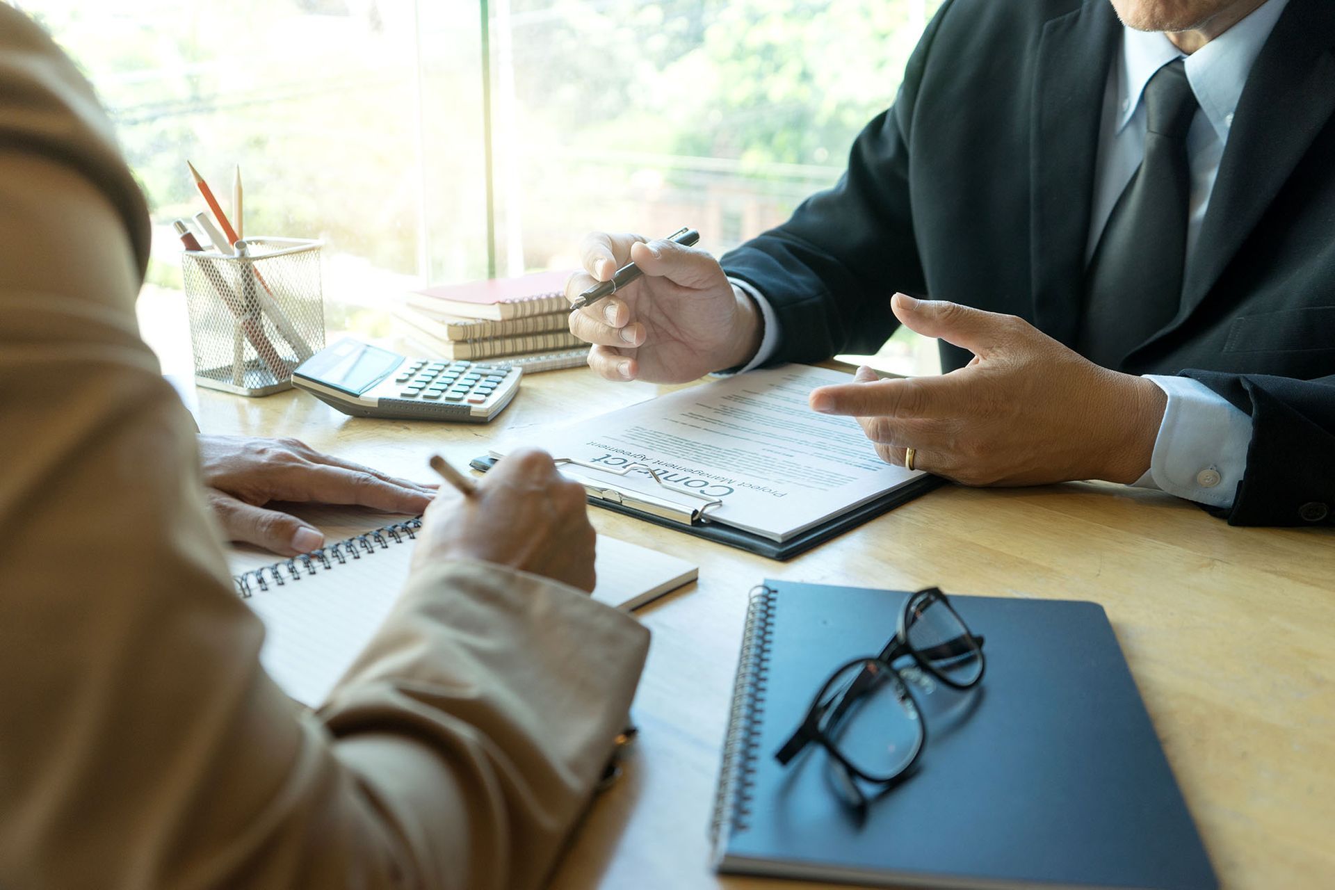 Two professionals in business attire confer at a desk with notebooks, a calculator, and glasses, reviewing a document.