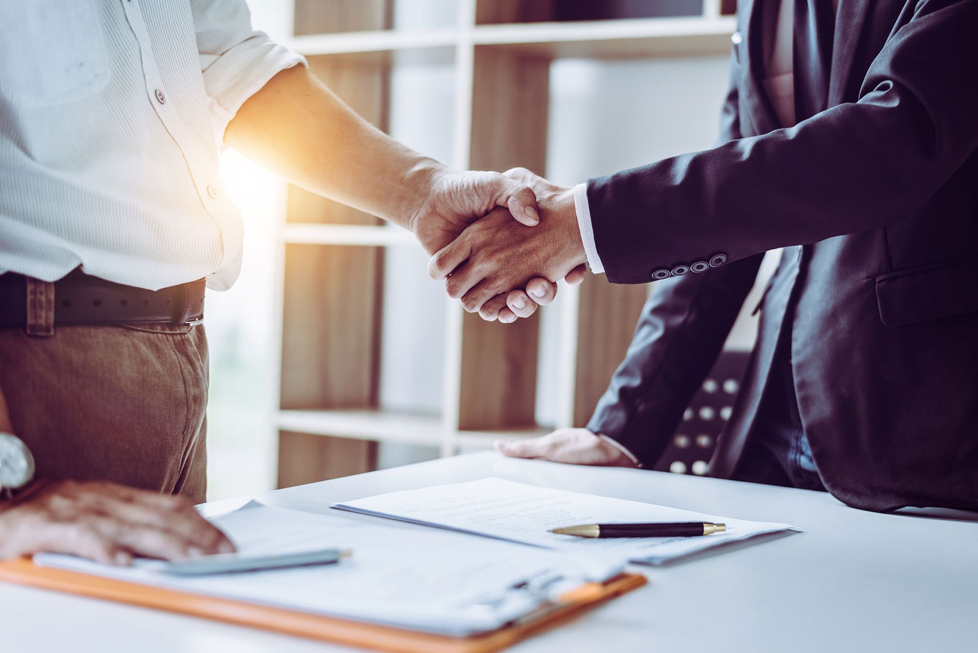 Two professionals in business attire shake hands over a desk with documents and a pen in a well-lit office setting.