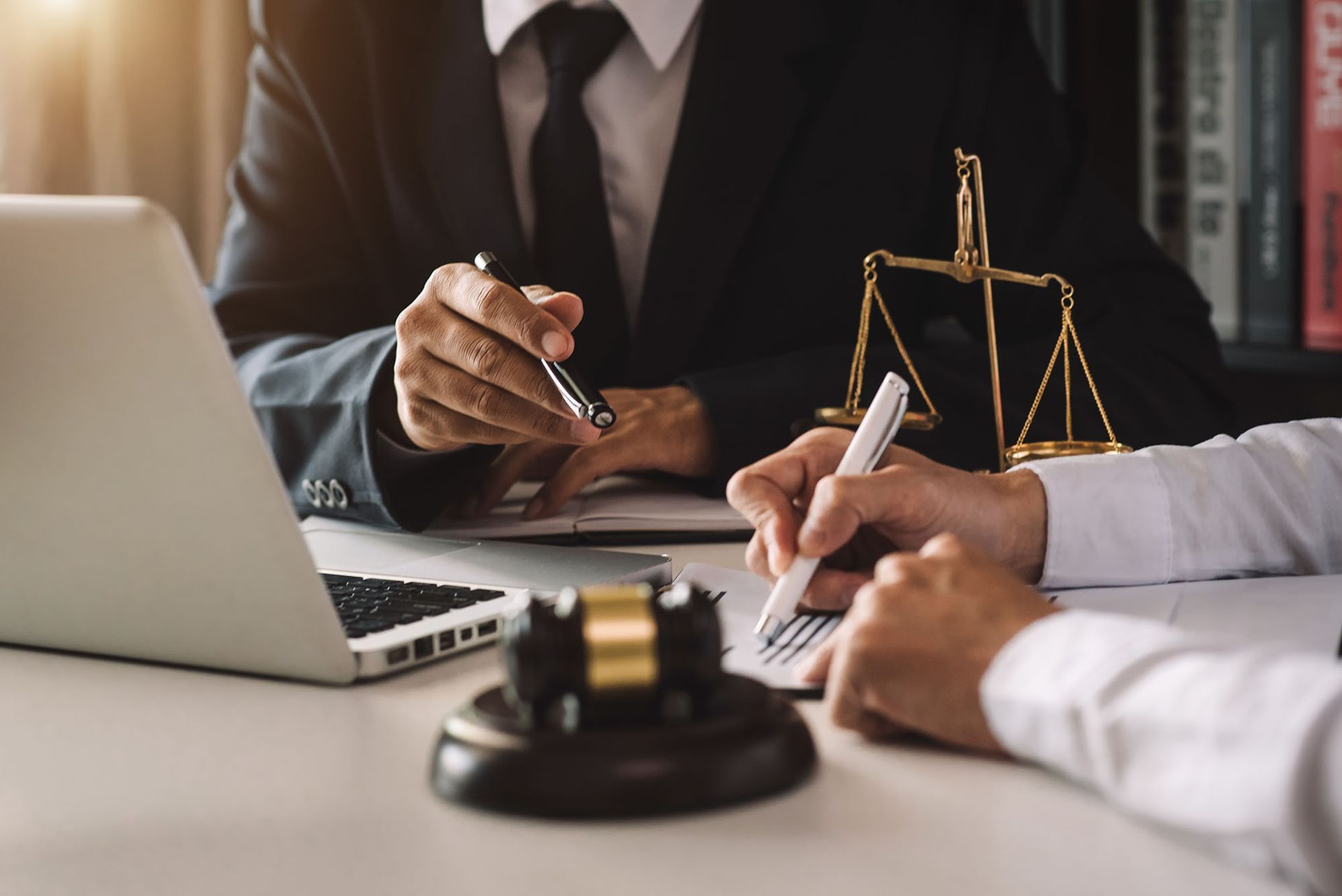 A legal professional and client review documents at a desk featuring a laptop, a gavel, and a set of scales.