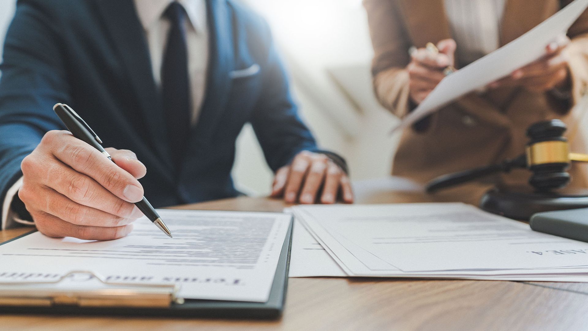 A person in a suit signs a legal document at a desk with another person and a gavel in the background.
