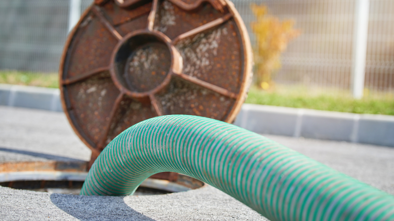 A green hose extends from an open manhole. A rusty, circular cover sits behind the opening.