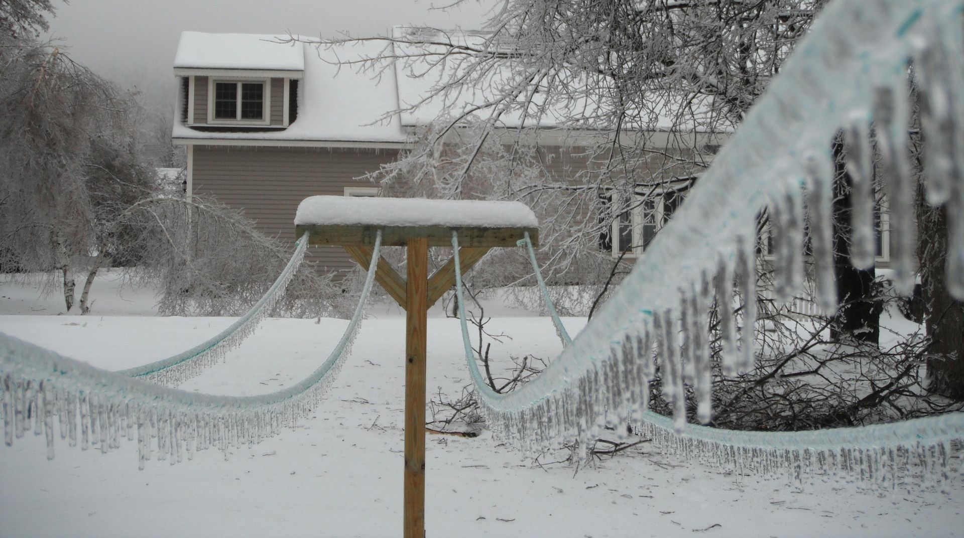 A snowy scene with a wooden bird feeder covered in snow and icicles, a house in the background, and ice-covered lines in the foreground.