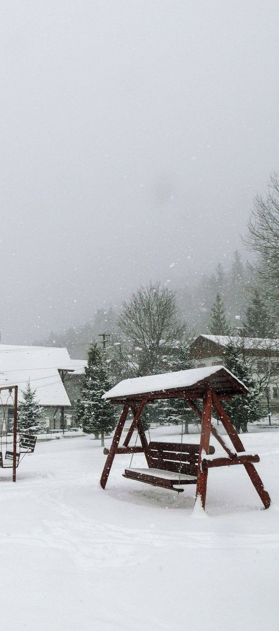A snow-covered yard with a wooden swing set. Snowflakes fall on the ground, a house, and trees.
