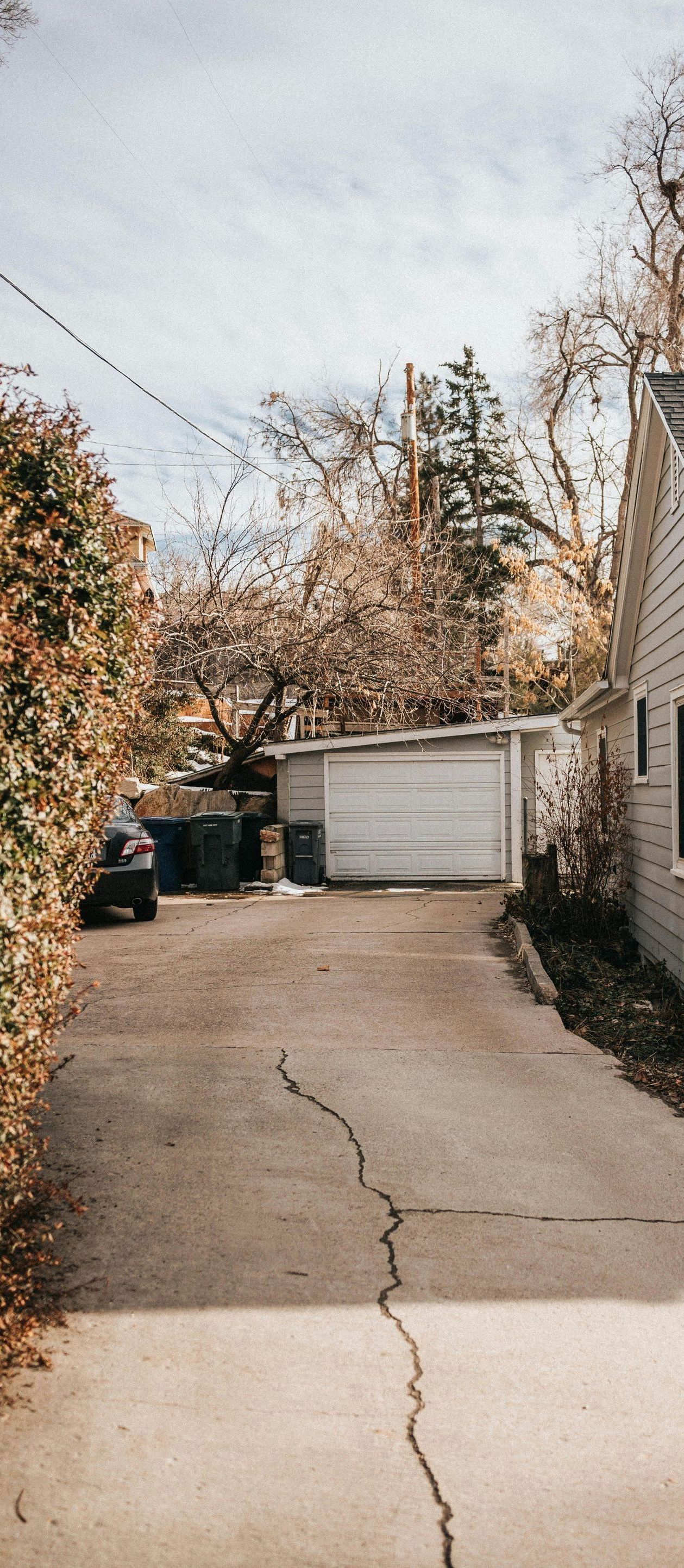 A driveway leading to a detached garage with a cracked concrete surface. Autumn trees flank the sides.