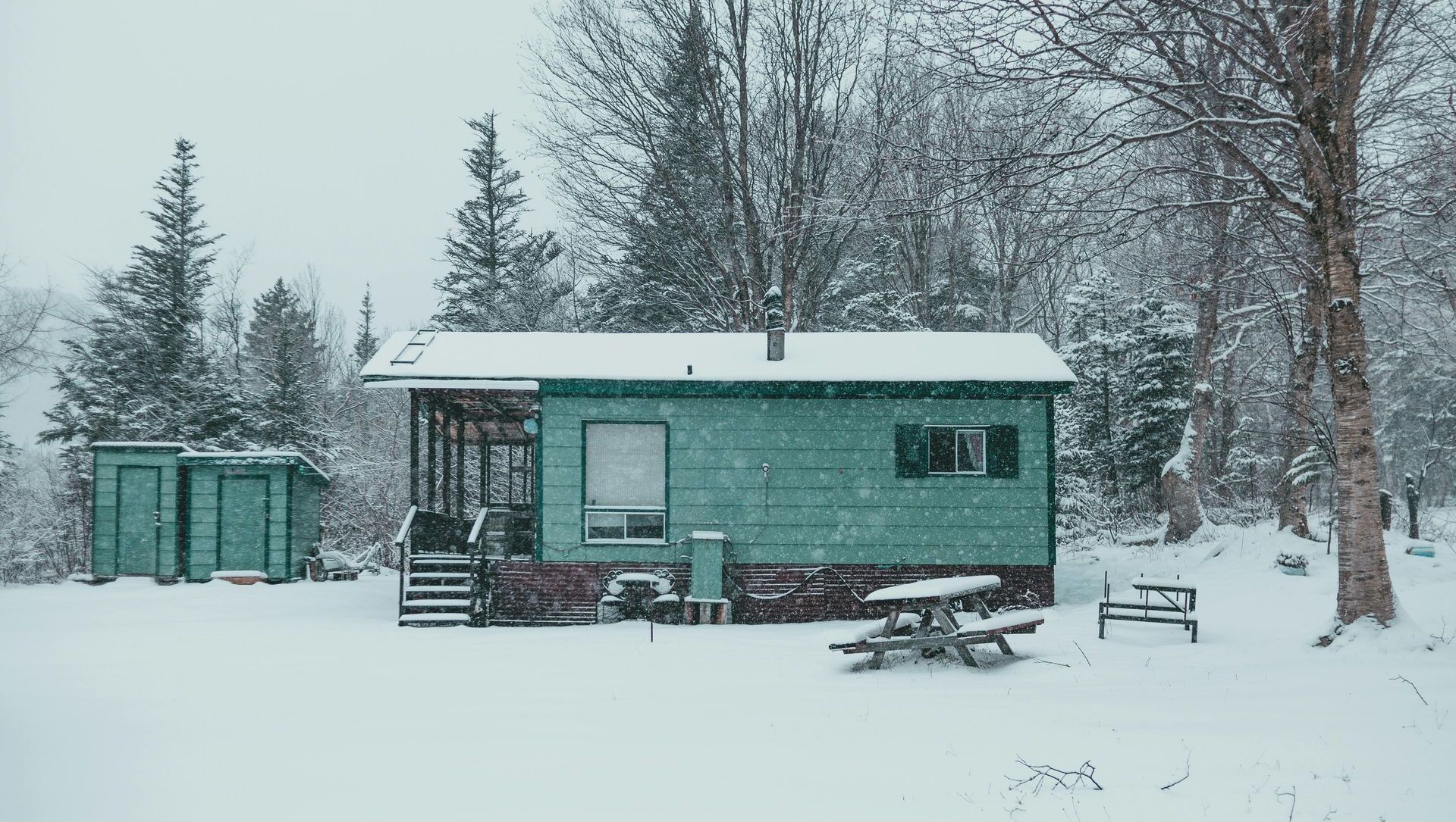 A teal cabin in a snowy landscape. Snow falls on the cabin, trees, and picnic table.