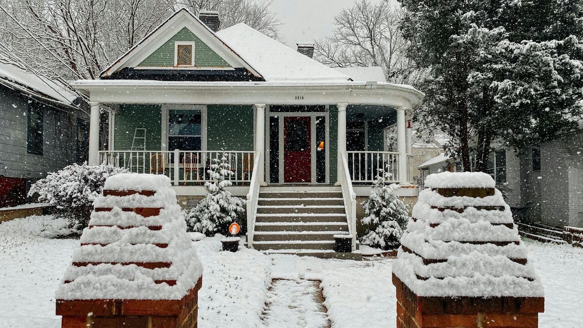 Snowy, two-story house with a green facade, red front door, and snow-covered porch. White snow falls on the lawn and brick pillars.