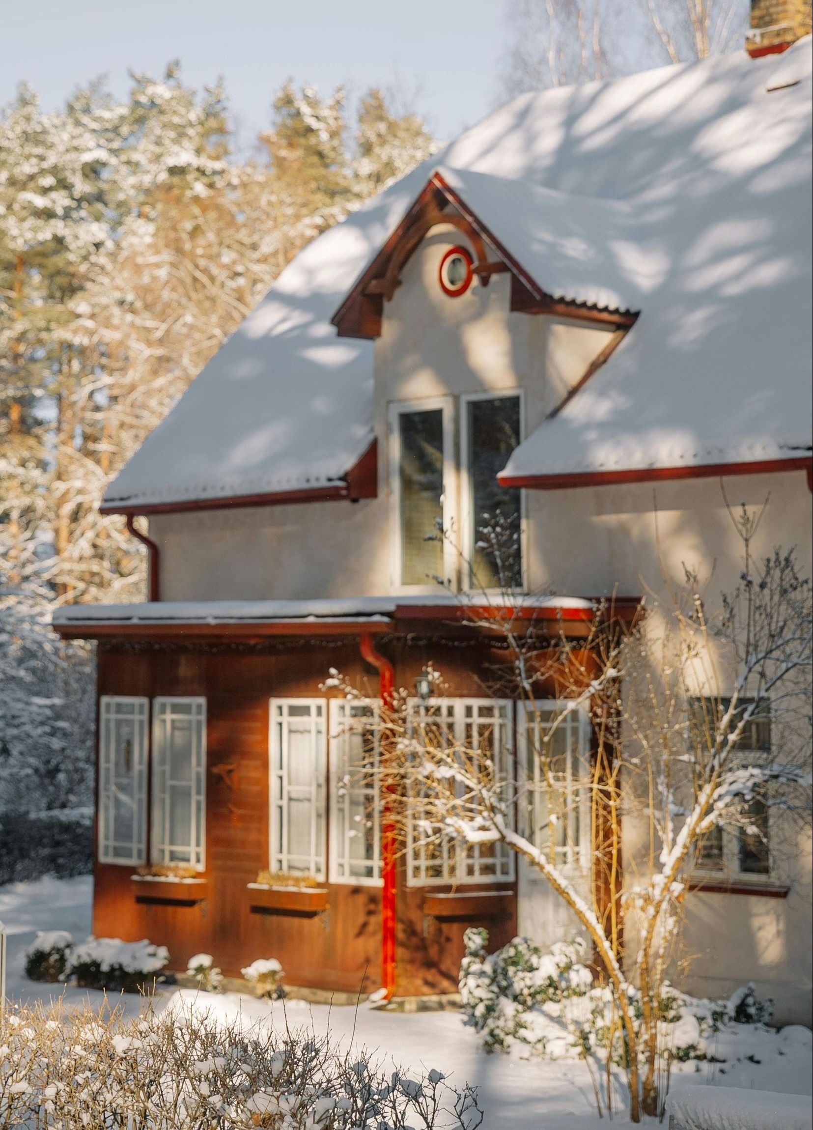 Snow-covered house with a wooden porch and red accents in a winter setting.  Trees and bushes are also covered in snow.