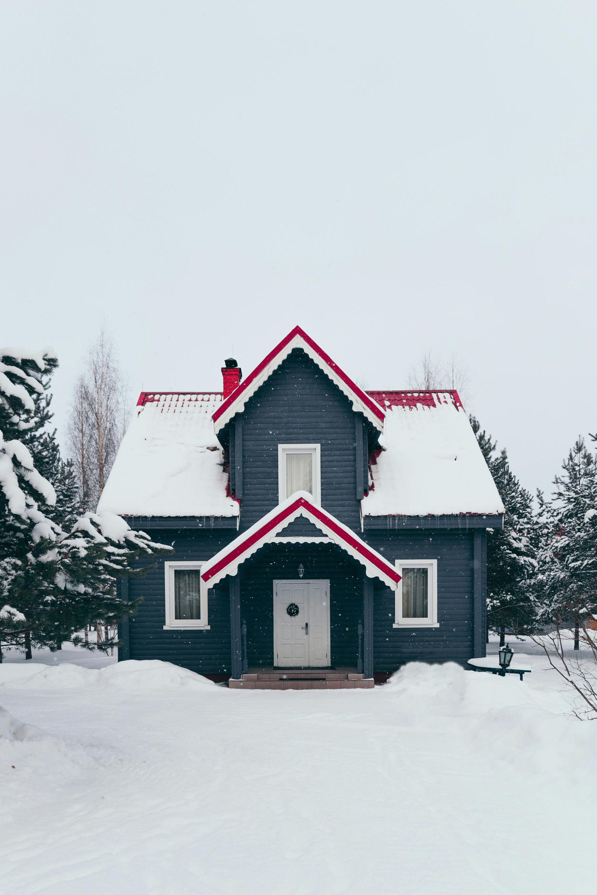 Snow-covered wooden house with red trim and roof. The gray house sits in a snowy landscape, surrounded by trees, with a white door.