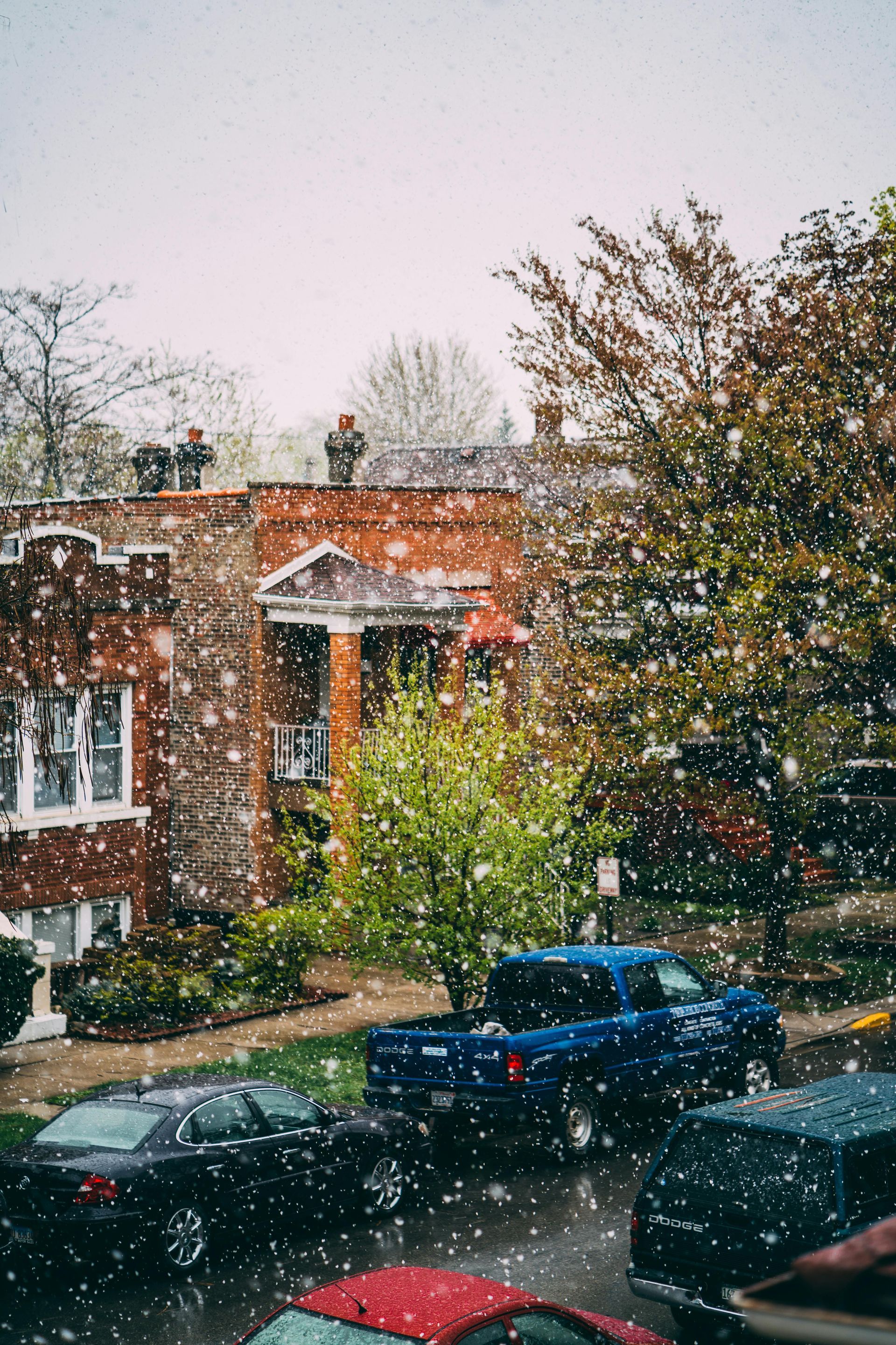 Snow falling on brick apartment building, cars parked on street, and two people on the roof.