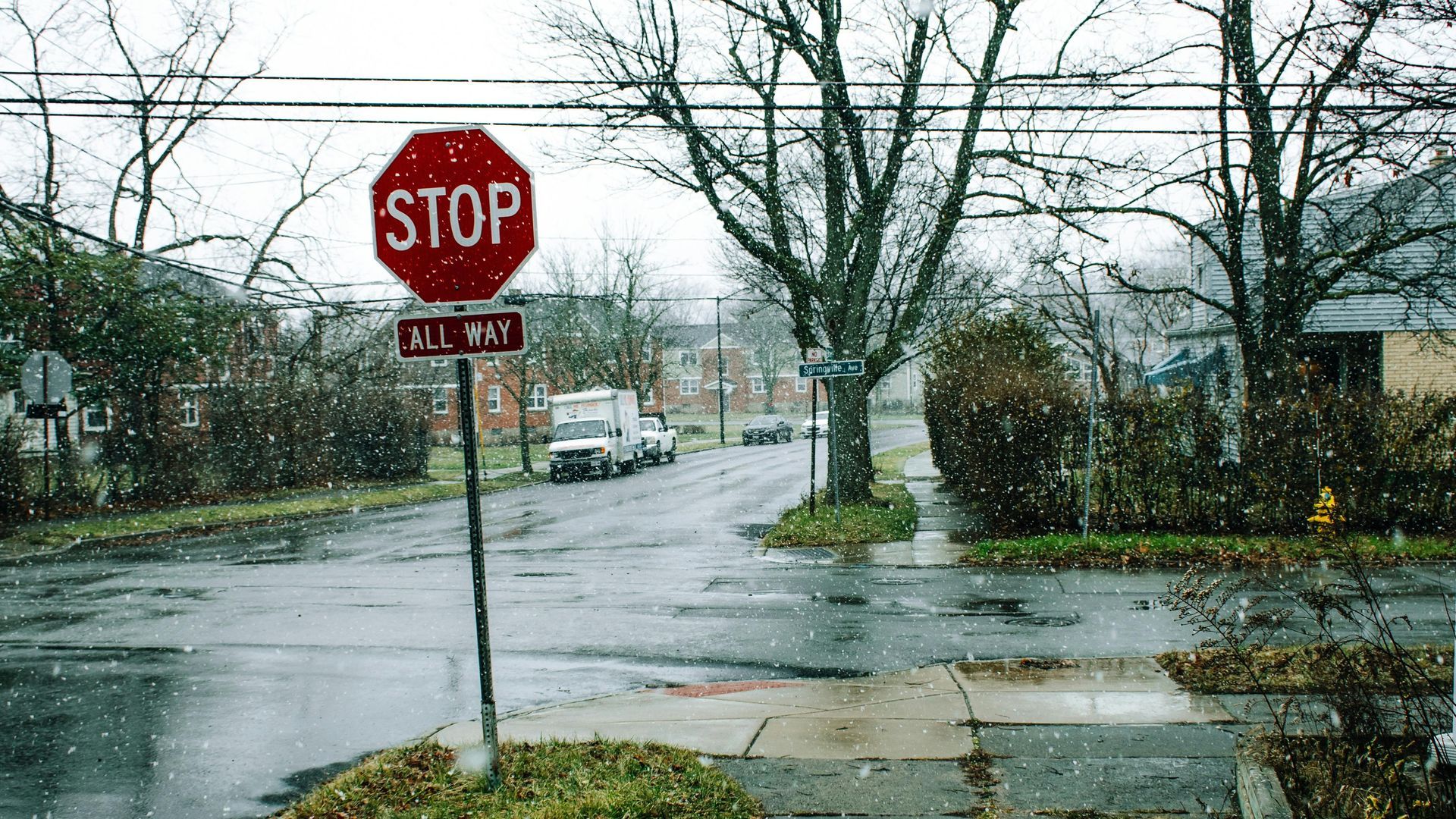 Stop sign at a wet intersection on a snowy day, houses and parked vehicles in the background.