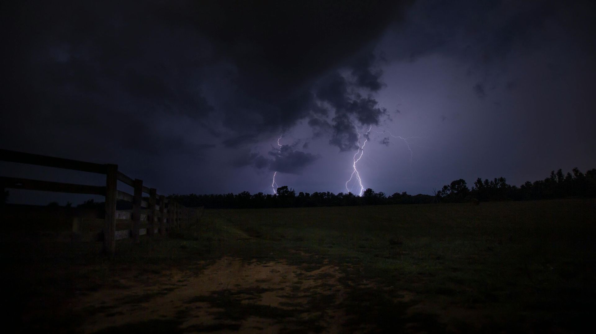 Lightning strikes over a field at night with a fence in the foreground.