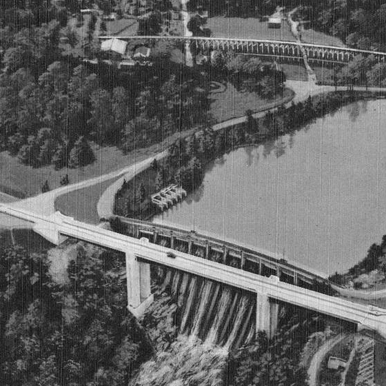 Aerial view of a dam and reservoir with water cascading down, bridge above, trees and buildings around.