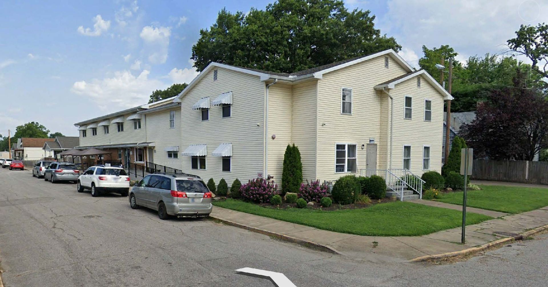 White apartment building with parked cars along a street and trees in the background