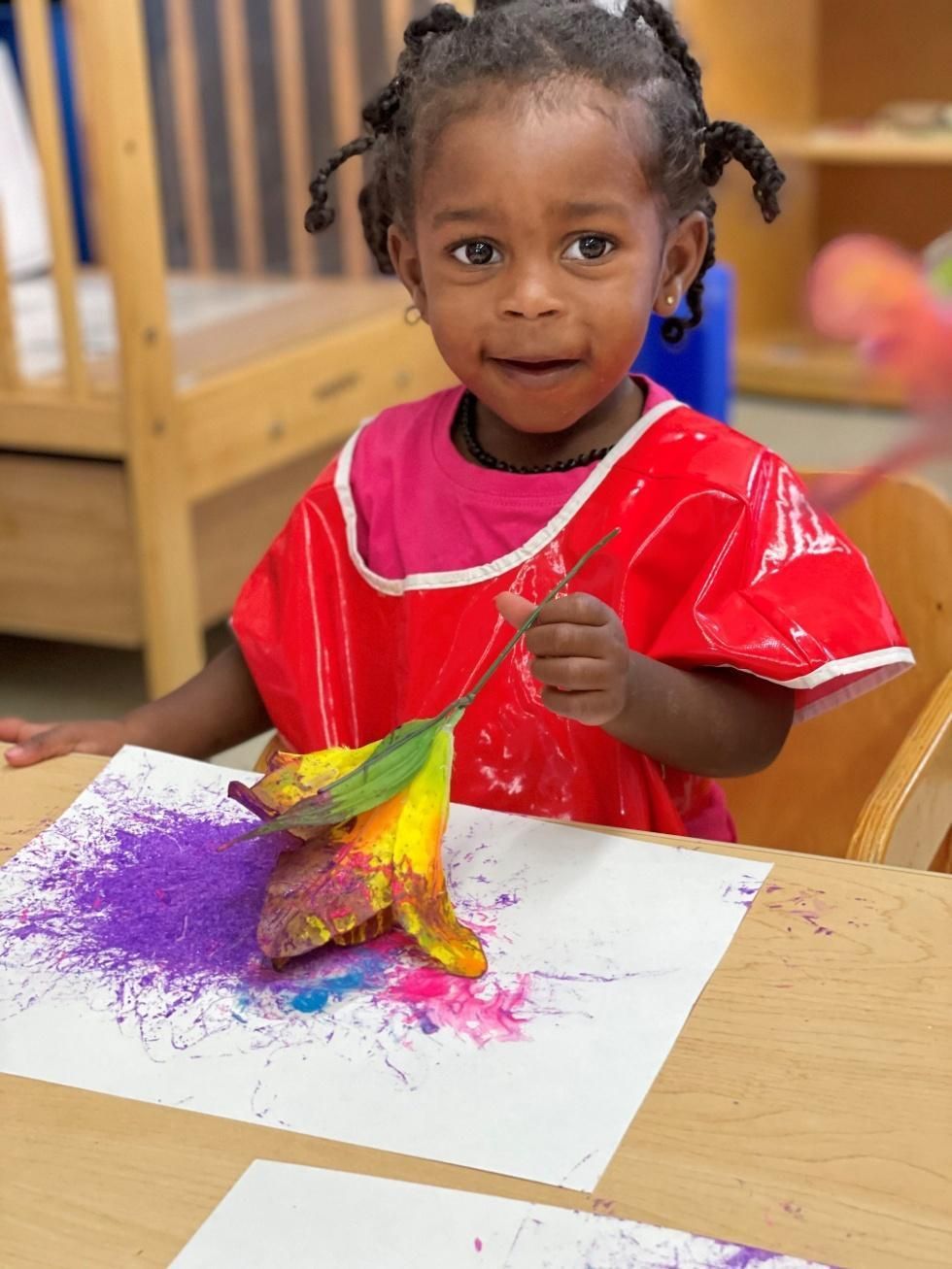 Child in a red smock painting with bright purple and yellow paint at a table.