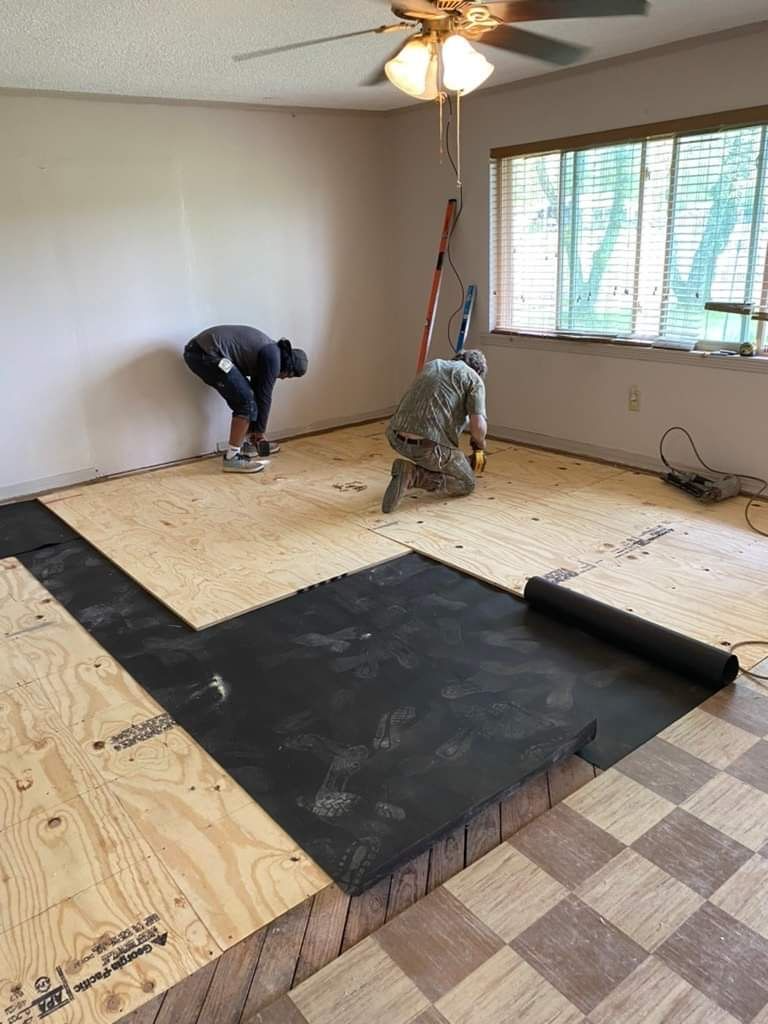 Two workers installing flooring in a room with a window. One kneels, the other stands, laying plywood.