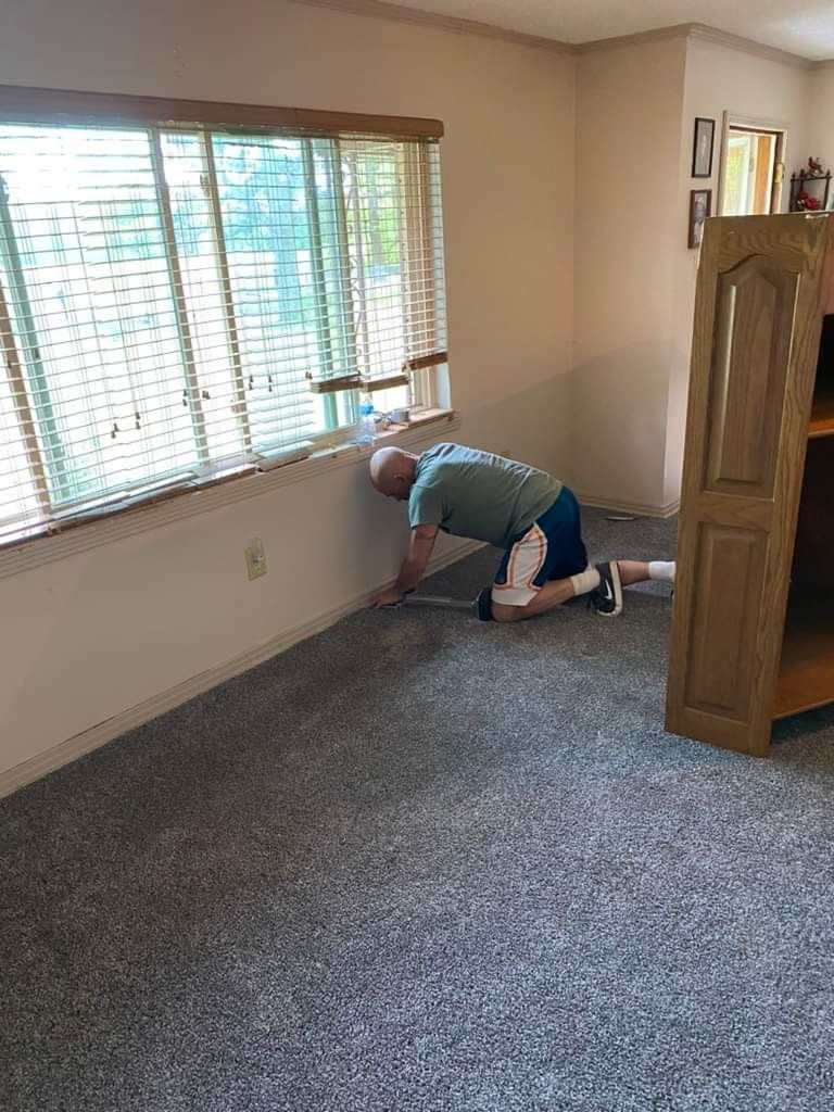 Man kneeling, installing grey carpet next to a window and wood bookcase.