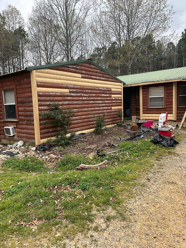 Log cabin being renovated; partially stripped wood siding. Green grass, trees, debris.