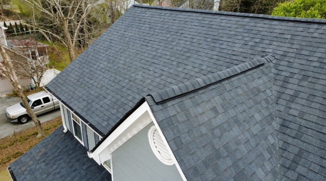 Aerial view of a dark gray shingled roof with dormer windows and a white house exterior.