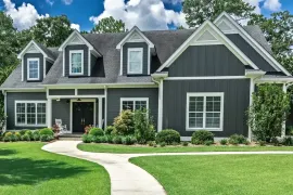Gray suburban house with white trim, front porch, manicured lawn, and curved walkway under a blue sky