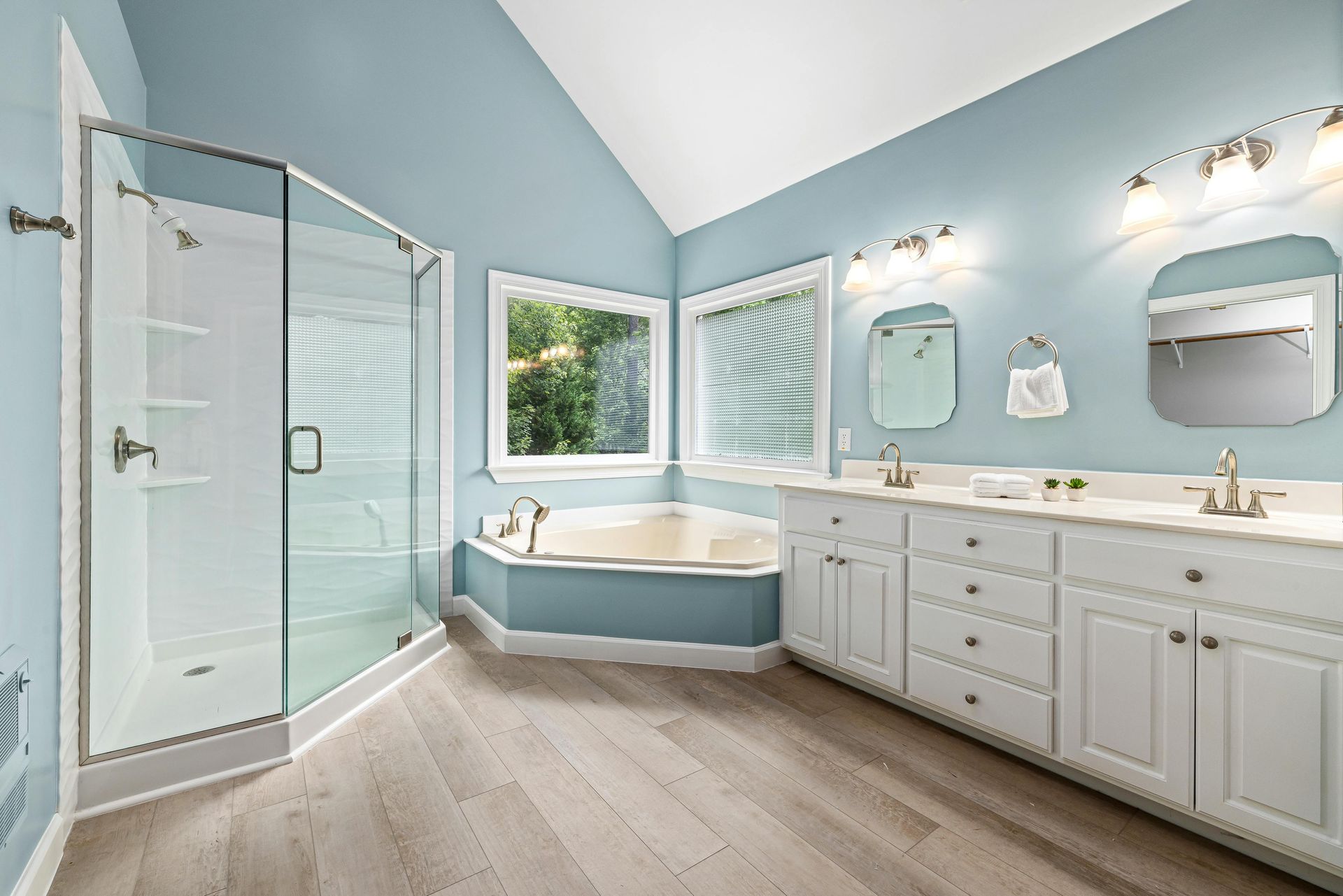 Bright blue bathroom with soaking tub, glass shower, double vanity, and two mirrors under skylights.