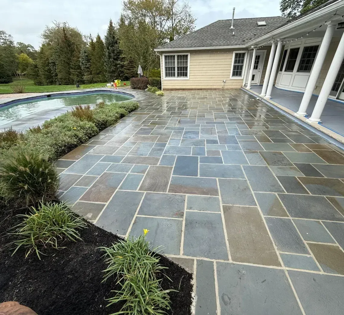 Stone patio beside a backyard pool and house, with gray-blue pavers and landscaped garden beds