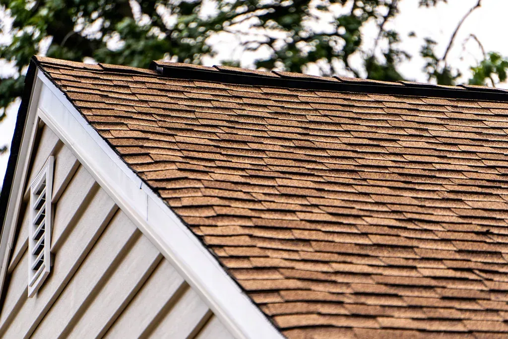 Close-up of a house roof with brown shingles and white siding beside trees