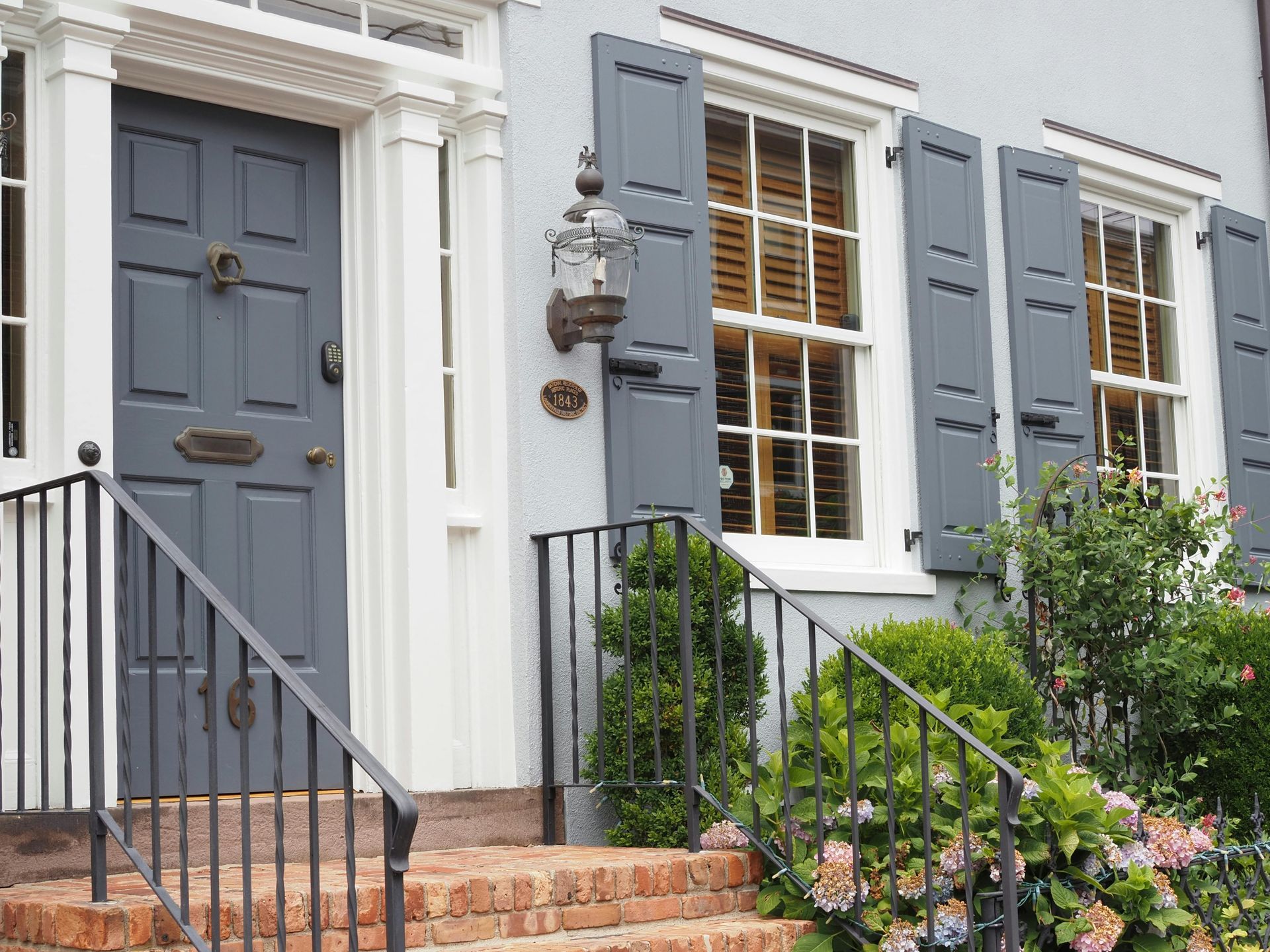 Gray front door with shutters, lantern, and garden beside brick steps on a white house facade