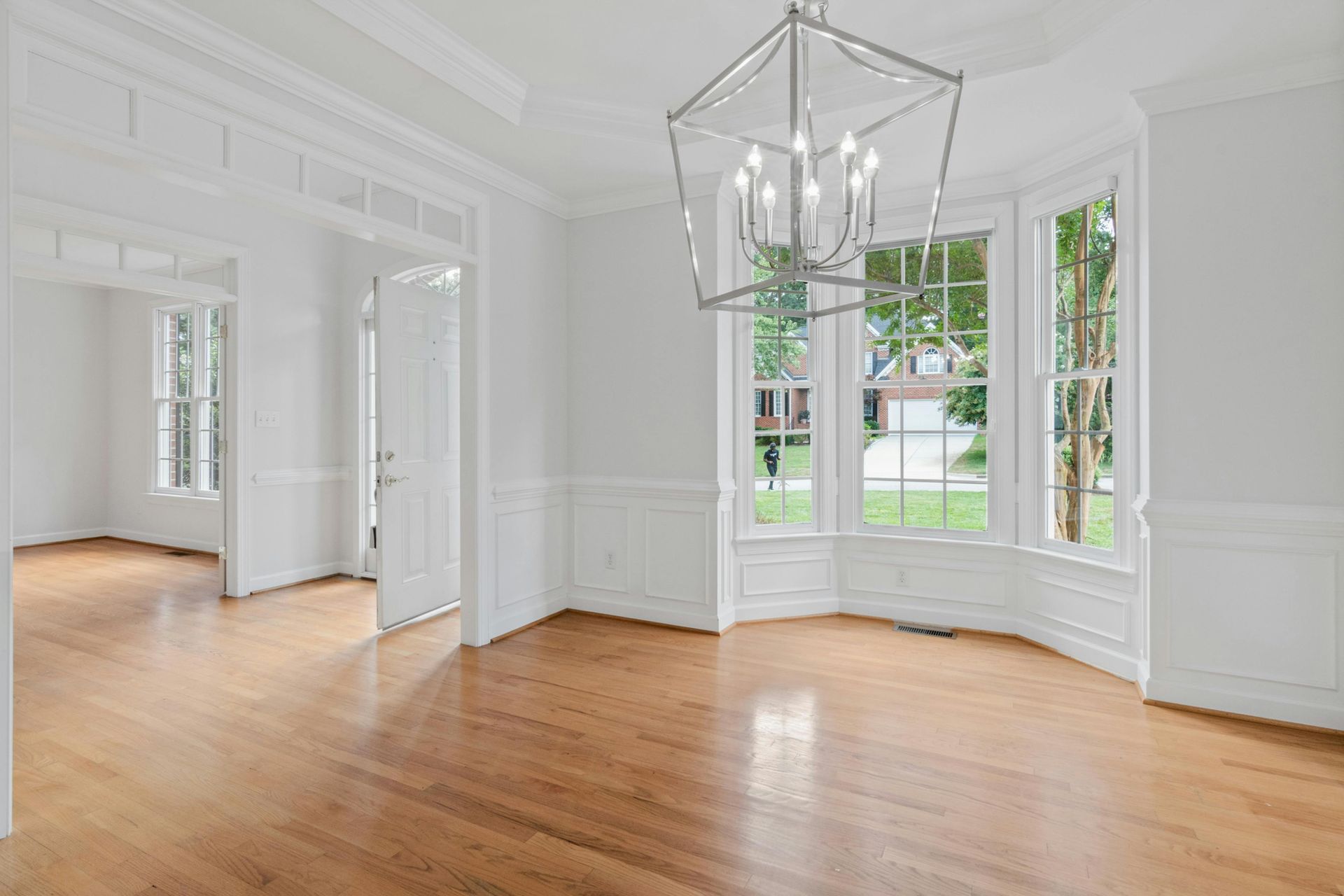 Bright empty dining room with white walls, hardwood floors, bay windows, and a modern chandelier.