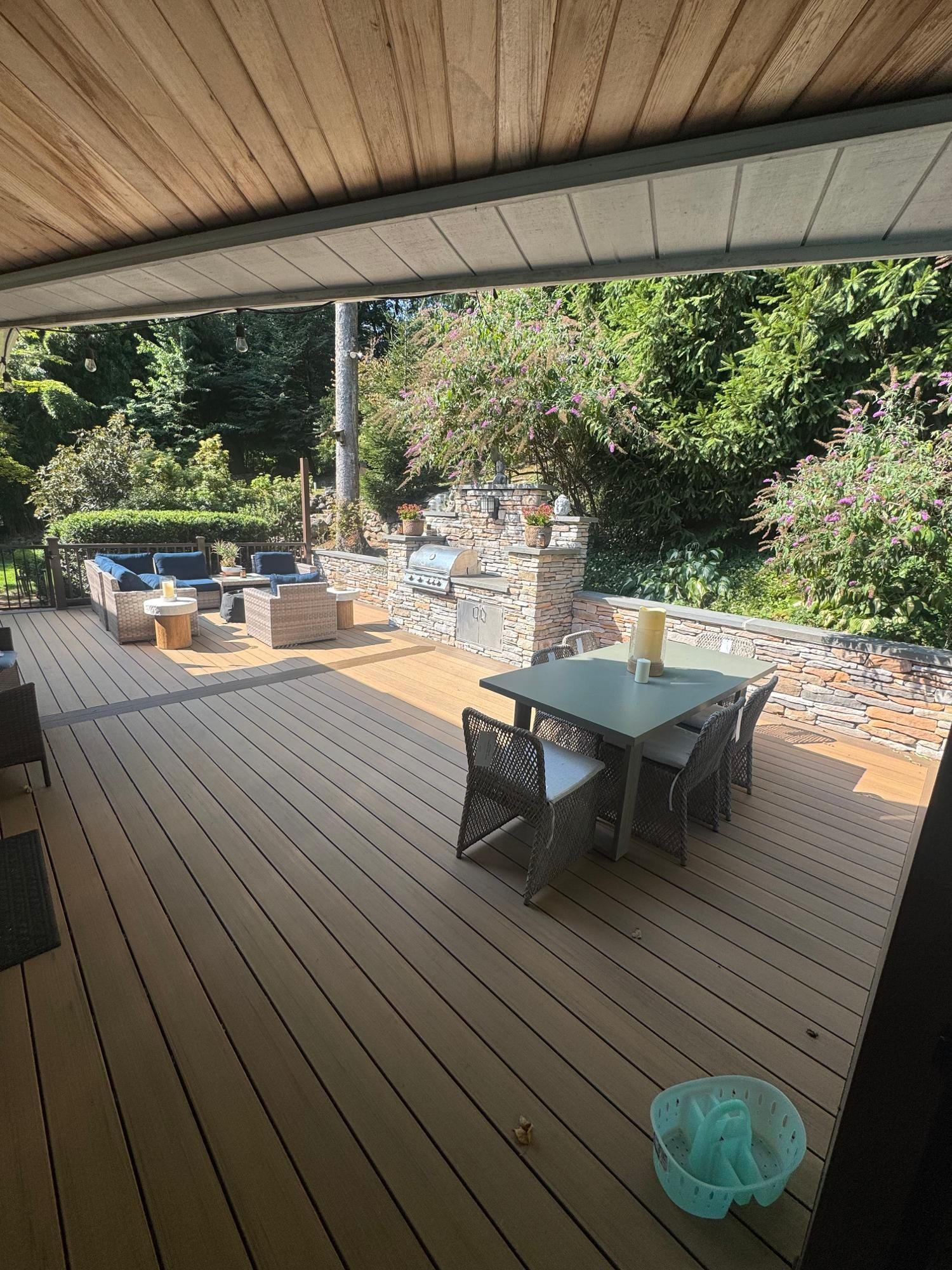 Covered wooden deck with outdoor dining table, chairs, grill, and garden plants in bright sunlight