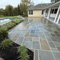 Stone patio beside a backyard pool and beige house, with garden beds and overcast sky.
