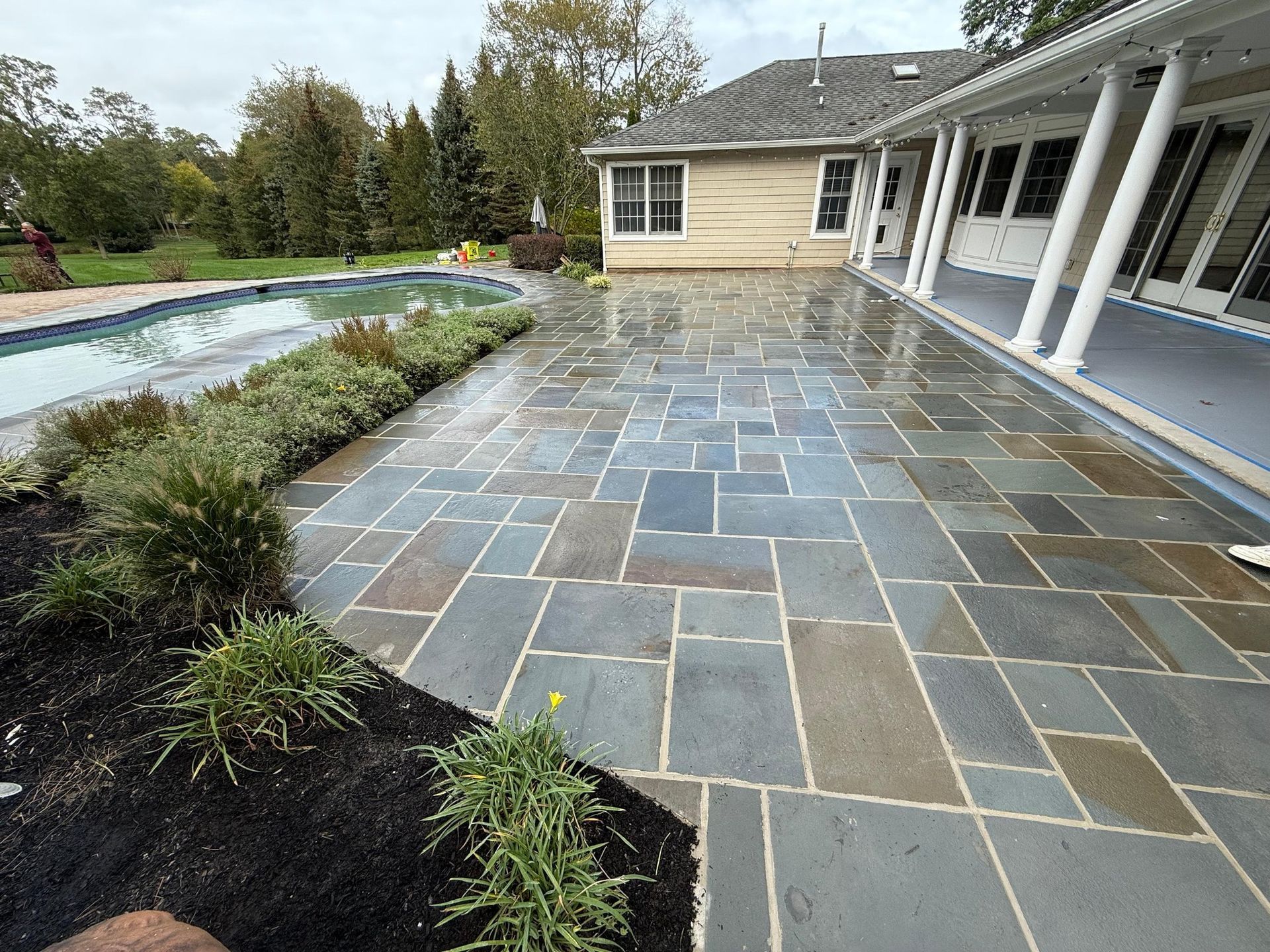 Stone patio beside a backyard pool and house, with gray-blue pavers and landscaped garden beds