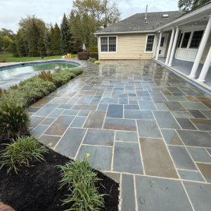 Stone patio beside a backyard pool and beige house, with garden beds and overcast sky.