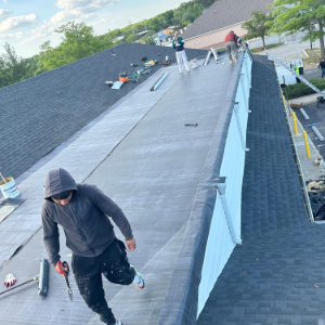 Workers on a rooftop, repairing dark shingles on a steep residential roof under a cloudy sky