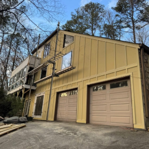 Tan-sided house with a double garage on a sloped wooded lot, showing exterior damage and exposed siding.