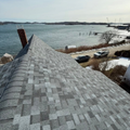 Snowy waterfront with a gray shingled roof or walkway leading to a calm harbor under a cloudy sky